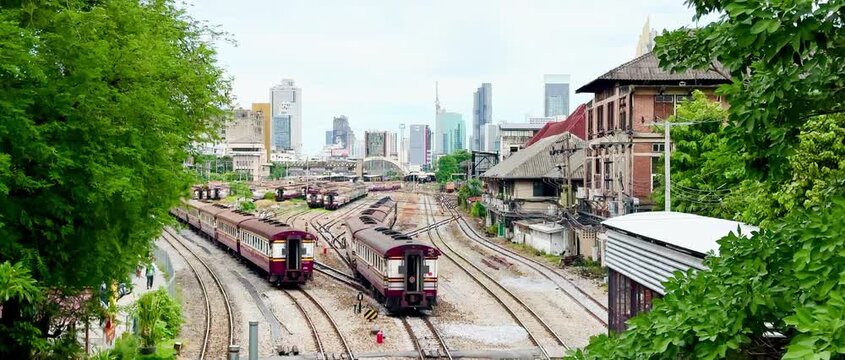 High-angle view of vintage passenger trains navigating multiple railway tracks at Hua Lamphong Station yard with the modern Bangkok city skyline and overcast sky in the background.