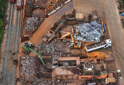 Aerial view of a scrap metal recycling facility with excavators sorting piles of metal, a truck being loaded, and railway tracks nearby in Unterwellenborn, Thuringia, Germany.