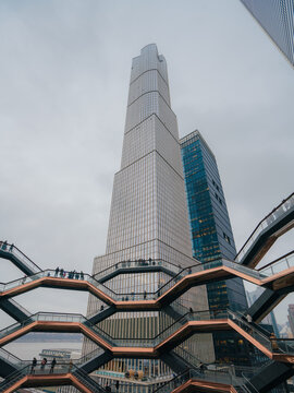 View of the Vessel honeycomb structure with modern skyscrapers in the background under a cloudy sky in New York, New York, United States.