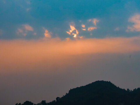 View of the sun setting behind the hills of Nhila in Teknaf, with soft orange light filtering through clouds over the landscape in Cox's Bazar, Chittagong Division, Bangladesh.