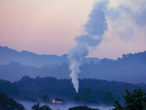 View of a thick plume of white smoke rising from a chimney against a backdrop of misty, layered hills at dawn in Nhila, Teknaf, Cox's Bazar, Chittagong Division, Bangladesh.
