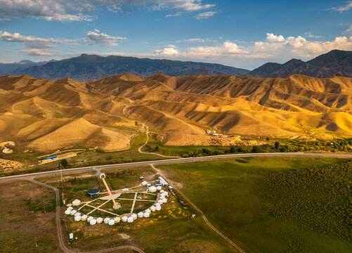 Aerial view of a traditional yurt camp arranged in a circle on a grassy plain, surrounded by arid rolling hills and mountains under a blue sky in Naryn, Naryn Region, Kyrgyzstan.