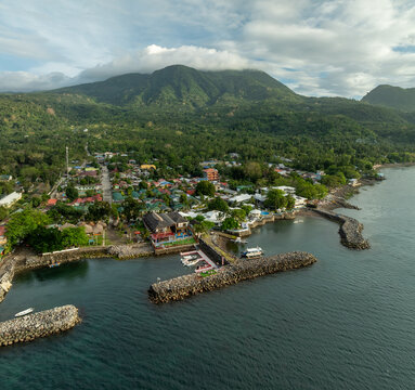 Aerial view of the coastal town of Mambajao with its breakwater, small boats, lush green mountains in the background, and clear blue sea in Mambajao, Camiguin, Philippines.