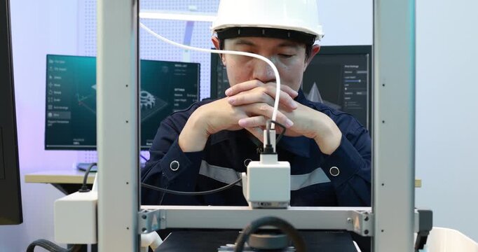 A technician in a hard hat looks thoughtfully at a machine in a modern lab or factory, with monitors and industrial equipment in the background.