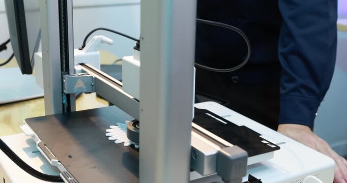 Person operating a 3d printer at work, printing a small white object on the build plate in a modern workshop.