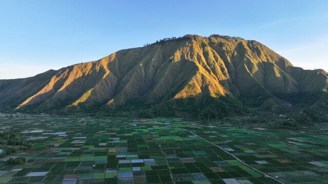 Aerial view of the Sembalun valley with its patchwork of agricultural fields and the slopes of Mount Rinjani illuminated by golden hour light in Sembalun, West Nusa Tenggara, Indonesia.