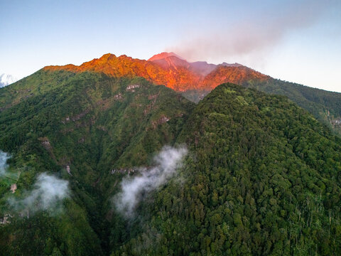 Aerial view of Mount Merapi with its lush green slopes and volcanic crater emitting smoke under the warm glow of sunrise in Selo, Central Java, Indonesia.