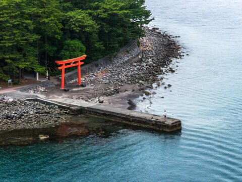 Aerial view of Mihama Beach featuring a vibrant red torii gate standing on the rocky shoreline with a lone fisherman on the concrete pier and lush green trees in Numazu, Shizuoka, Japan.