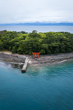 Aerial view of Osezaki Torii gate standing on the rocky shore of Mihama Beach, surrounded by lush green trees and the calm blue waters of Suruga Bay, Numazu, Shizuoka, Japan.