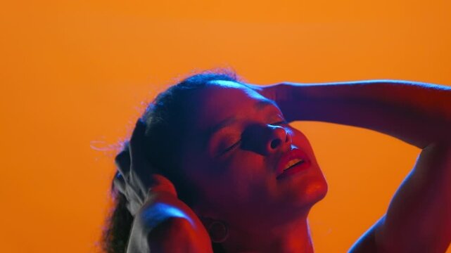 Close shot of young latin american woman with curly hair illuminated with red and blue led touches her face and hair with pleasure on a solid orange background | Perfume commercial concept