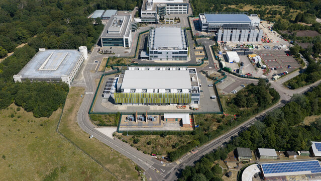 Aerial view of the Kao Data campus featuring modern industrial buildings, data center facilities, and surrounding green woodland in Harlow, England, United Kingdom.