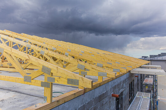 View of wooden roof trusses under construction on a concrete building with scaffolding against a dark stormy sky in Latvia.
