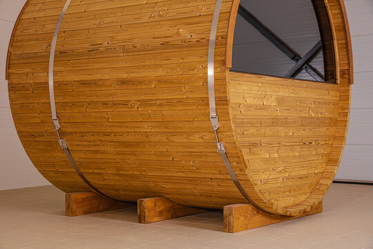View of a modern wooden barrel sauna with horizontal planks, metal tension straps, and a circular window, resting on wooden supports on a tiled floor in Latvia.