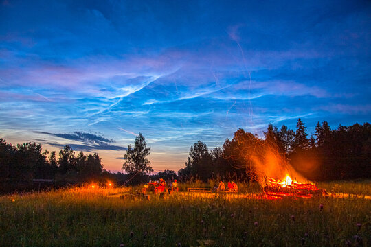 View of a bonfire burning in a field at night with people sitting around it under a sky filled with noctilucent clouds and silhouettes of trees in Latvia.