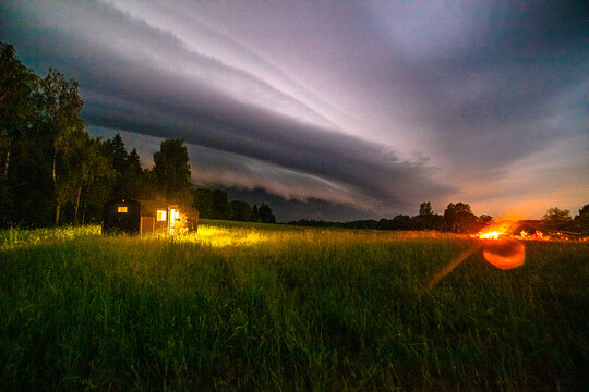 View of a small illuminated cabin in a grassy field under a dramatic stormy sky with dark rolling clouds at night in Latvia.