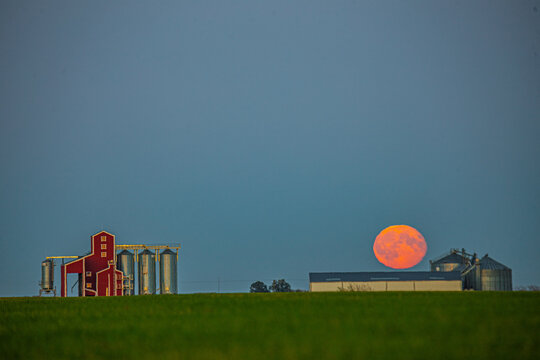 View of a red farm building and silver grain silos set against a large orange moon rising over a green field in Latvia.