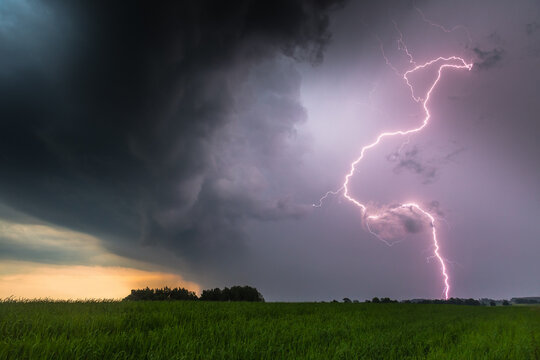 View of a powerful lightning strike illuminating the dark, dramatic storm clouds over a vibrant green field in Latvia.