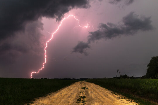 View of a dramatic lightning strike illuminating the dark storm clouds above a rural dirt road winding through a grassy field in Latvia.