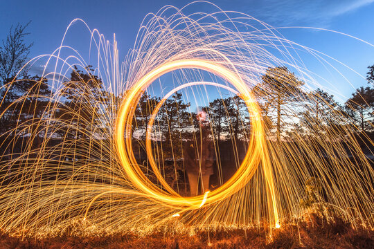 View of a person creating circular light trails with burning steel wool in a forest at dusk in Latvia.