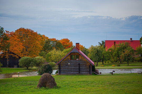 View of a rustic log cabin with a red roof situated by a tranquil pond surrounded by vibrant autumn trees and a hay bale in a grassy field in Latvia.