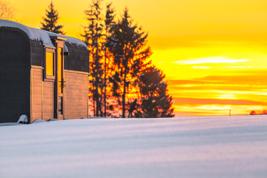 View of a modern tiny house cabin situated in a snow-covered field during a vibrant golden hour sunset with silhouetted pine trees in the background in Latvia.