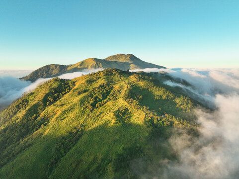 Aerial view of Gunung Mongkrang with lush green mountain slopes and rolling hills surrounded by thick clouds under a clear blue sky in Jatiyoso, Central Java, Indonesia.