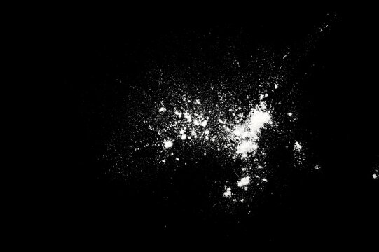 White flour powder splash on a black background with scattered particles and small bursts, creating a dark abstract texture with strong contrast.