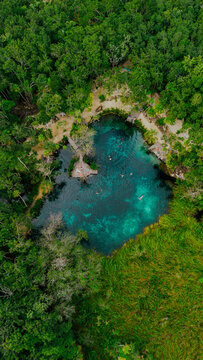 Aerial view of Cenote Corazon del Paraiso surrounded by lush jungle with turquoise water, wooden platforms, and people swimming in Tulum, Quintana Roo, Mexico.