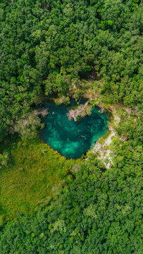 Aerial view of Cenote Corazon del Paraiso surrounded by dense tropical jungle with turquoise water and people swimming in Tulum, Quintana Roo, Mexico.