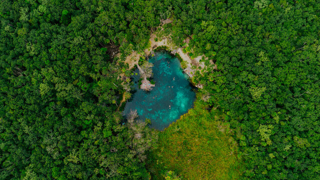 Aerial view of Cenote Corazon del Paraiso surrounded by dense tropical jungle with people swimming in the clear turquoise water in Tulum, Quintana Roo, Mexico.