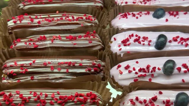 Close-up assorted eclairs in cafe display. Glossy chocolate and pastel pink glazes topped with red crumble and single blueberry