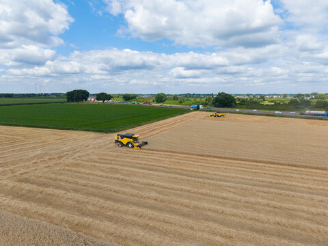 Aerial view of a yellow combine harvester working in a golden wheat field with another harvester visible in the distance and a road with traffic in the Netherlands.
