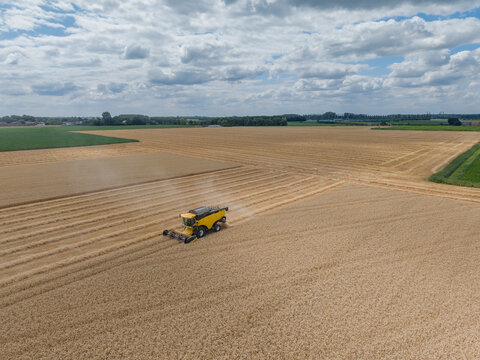 Aerial view of a yellow combine harvester working in a vast golden wheat field under a cloudy blue sky in the Netherlands.
