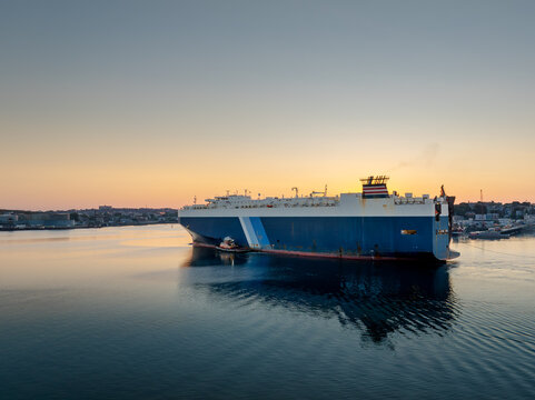 Aerial view of a large vehicle carrier ship being assisted by a tugboat in the harbor at sunrise with the city skyline in the background in Boston, Massachusetts, United States.