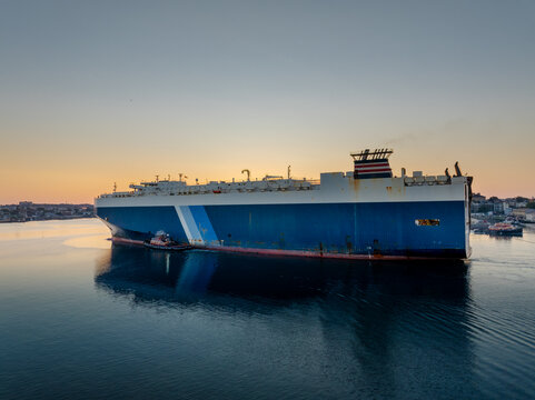 Aerial view of a large vehicle carrier ship coming into a harbor at sunrise, accompanied by a small tugboat on the calm water. Boston, Massachusetts, United States.