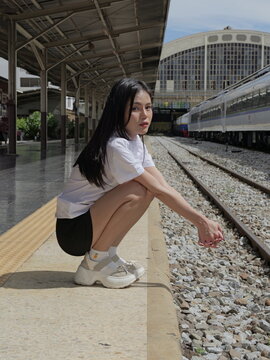 A young Asian woman wearing a university student uniform crouches on the edge of an open-air train platform