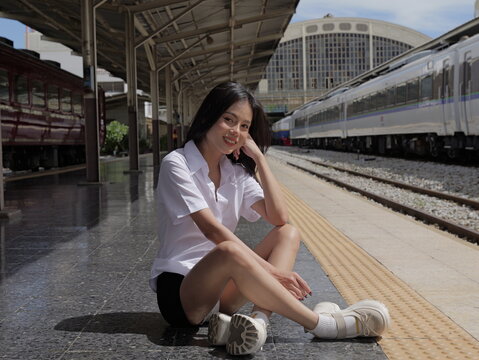 A young smiling Asian woman wearing a university student uniform sits cross-legged on a train station platform