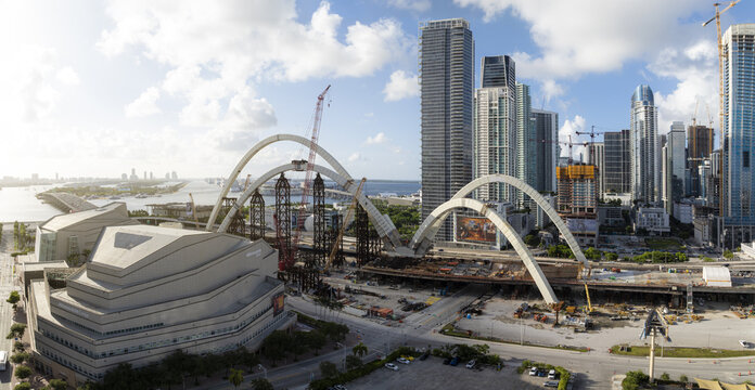 Aerial view of the Signature Bridge under construction with large white arches, cranes, and the downtown Miami skyline in the background, Miami, Florida, United States.