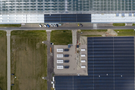 Aerial view of a large-scale solar farm featuring rows of photovoltaic panels and energy storage battery containers adjacent to a greenhouse facility in the Netherlands.