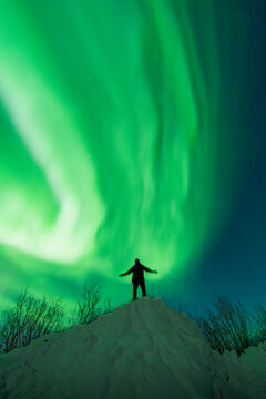 View of a person standing on a snow-covered hill with arms outstretched under the vibrant green aurora borealis in the night sky in Tromso, Troms, Norway.