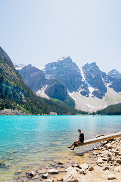 View of a man sitting on a log extending into the turquoise water of Moraine Lake with snow-capped mountains in the background in Improvement District No. 9, Alberta, Canada.