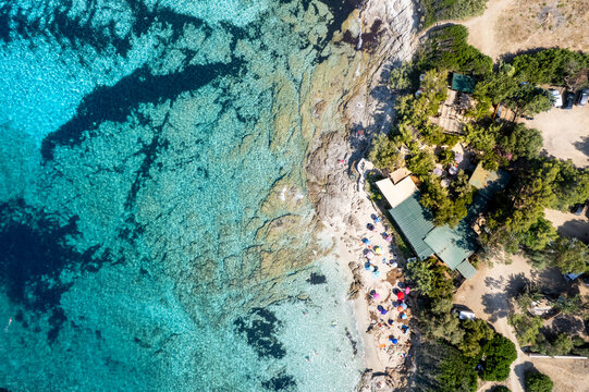 Aerial view of Plage de Ghjunchitu showing the turquoise waters, rocky coastline, sandy beach with colorful umbrellas, and surrounding greenery in Corbara, Corse, France.