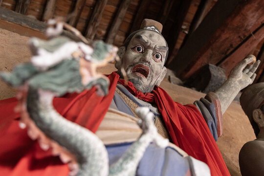 View of a traditional painted statue with a red robe and a dragon figure in the foreground, set against wooden architectural beams in Yuci Ancient Town, Yuci, Shanxi, China.