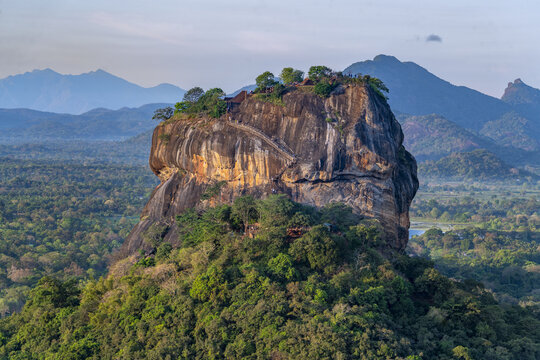 View of Sigiriya Lion Rock rising above the lush green jungle canopy with ancient stone staircases carved into the rock face in Sigiriya, Central Province, Sri Lanka.