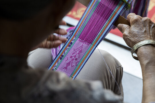 View of a woman carefully weaving intricate Nushu script patterns into a vibrant purple and multi-colored textile, showcasing traditional craftsmanship in Yongzhou, Hunan, China.