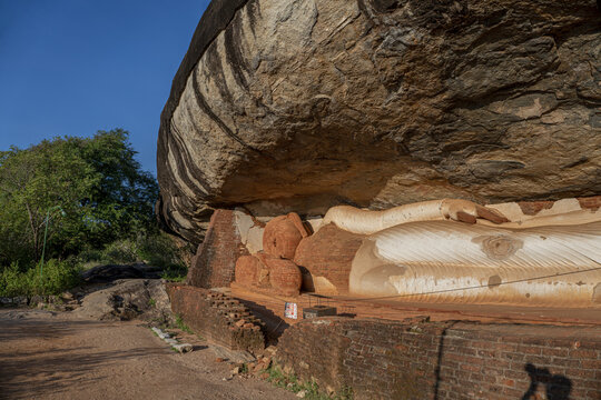 View of the ancient reclining Buddha statue at Pidurangala temple, featuring weathered brickwork and natural stone overhang under a clear blue sky in Sigiriya, Central Province, Sri Lanka.
