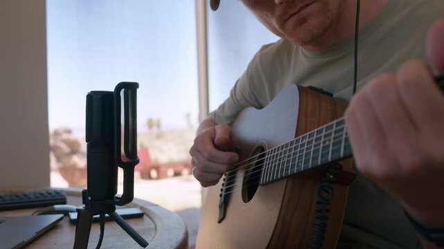 A close-up of a man recording acoustic guitar with a studio microphone beside a desert window.