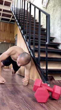 Slow motion vertical shot of a shirtless man doing knuckle push-ups on a wooden floor at home. Red dumbbells sit in the foreground. Great for fitness, strength training and home workout concepts.