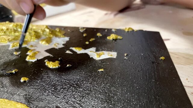 Slow motion macro close up of an artist's hand using a fine brush to paint black details on a heavily textured gold canvas. Modern abstract mixed media fine art creation process.