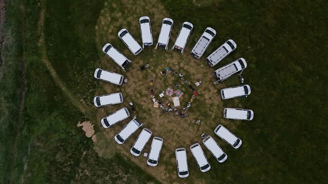 Aerial view of a circle of white camper vans parked on a grassy field with people gathering in the center in Iceland.